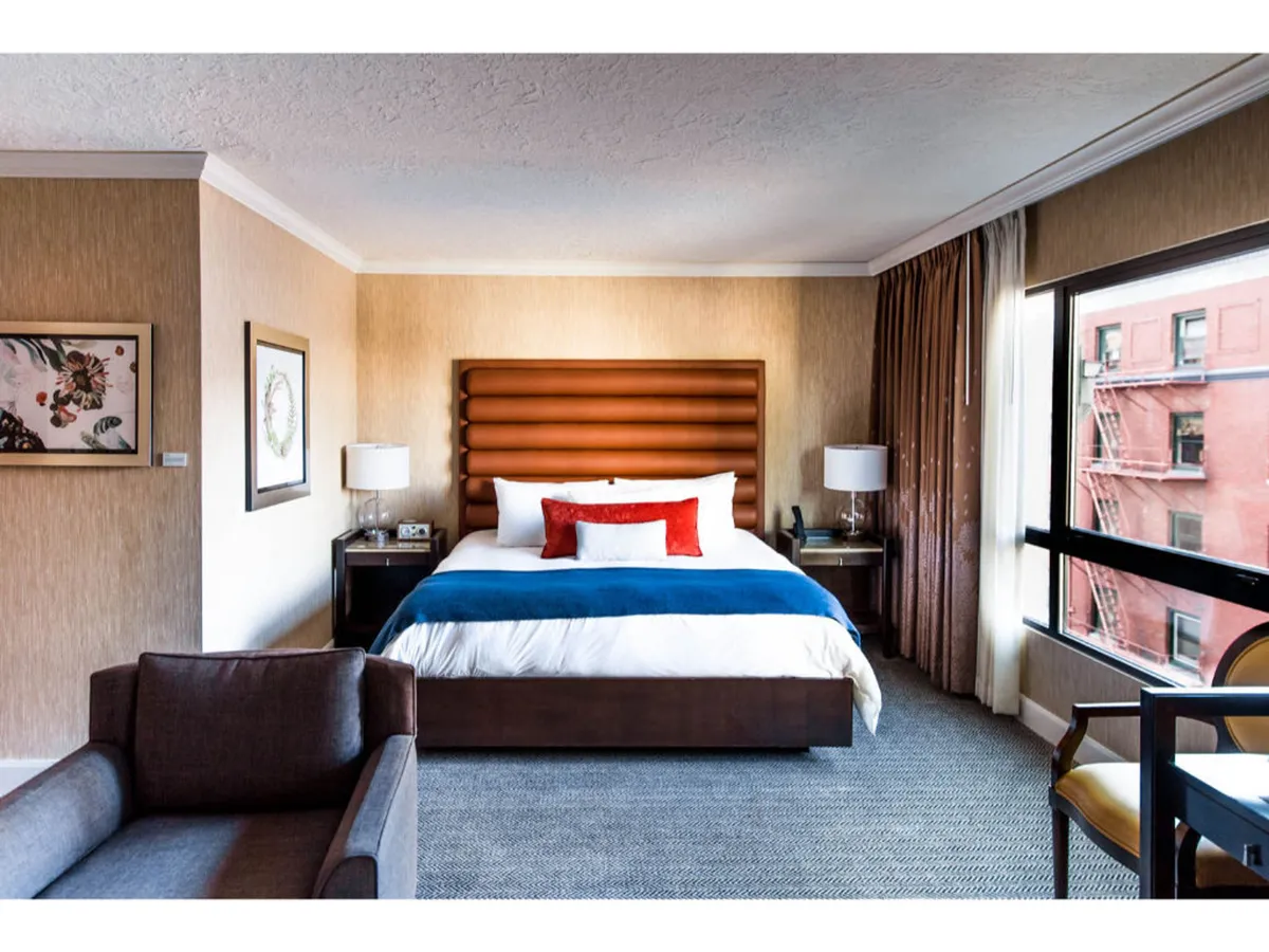 A spacious studio bedroom area featuring a king bed with an upholstered headboard, layered with decorative pillows and crisp linens. Sunlight streams in from a large window, illuminating locally inspired artwork above the bed and offering a refreshing view. The surrounding finishes are contemporary and high quality, with a plush ambiance inviting relaxation.