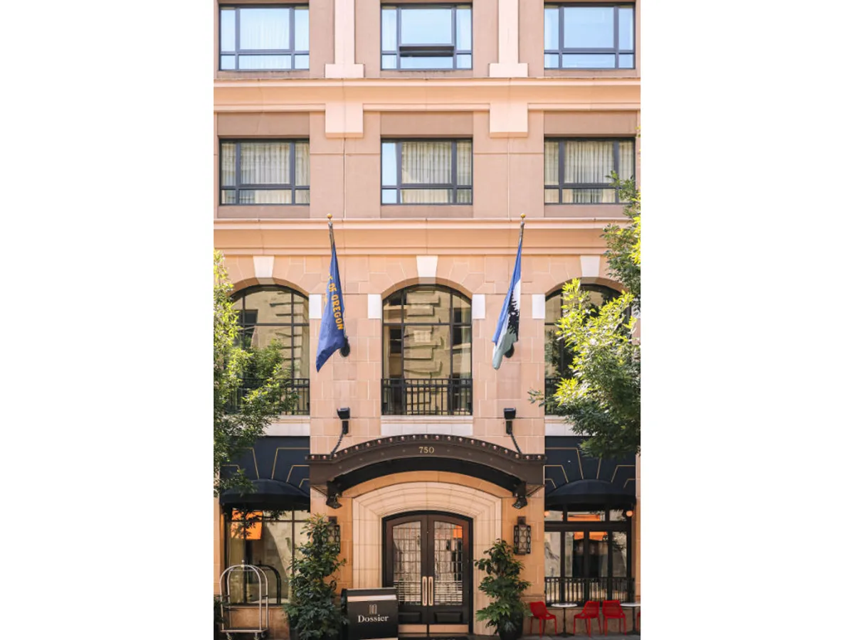 An exterior view of the main entrance with flags, prominent windows, and landscaped surroundings. The inviting facade hints at luxury and comfort, while people at the entrance suggest a welcoming sense of community and arrival.