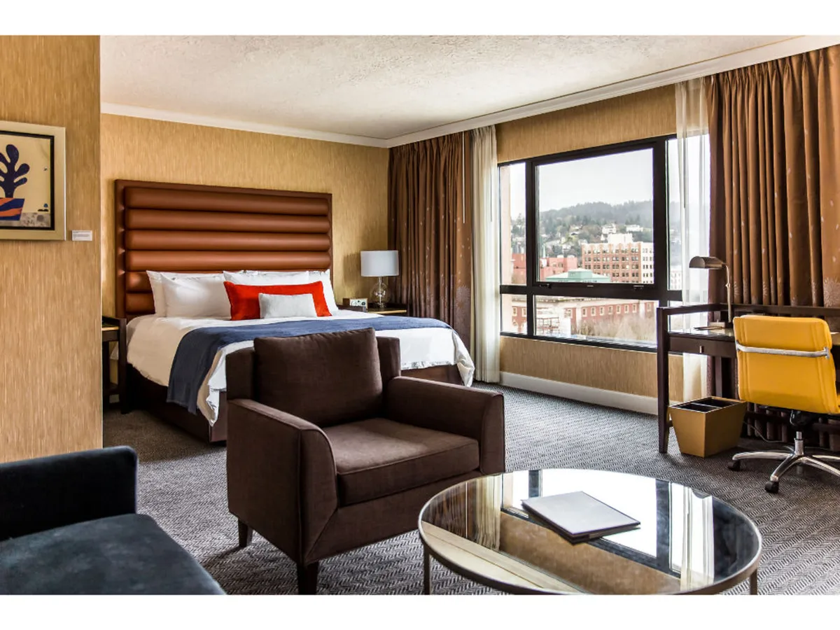 The image captures a chic loft suite featuring a plush bed with an elegant headboard, a large window showcasing sweeping city views of Portland, a cozy seating area perfect for unwinding, and a modern work desk bathed in natural light. The space is designed with stylish decor and thoughtfully chosen amenities, creating a tranquil yet sophisticated atmosphere.