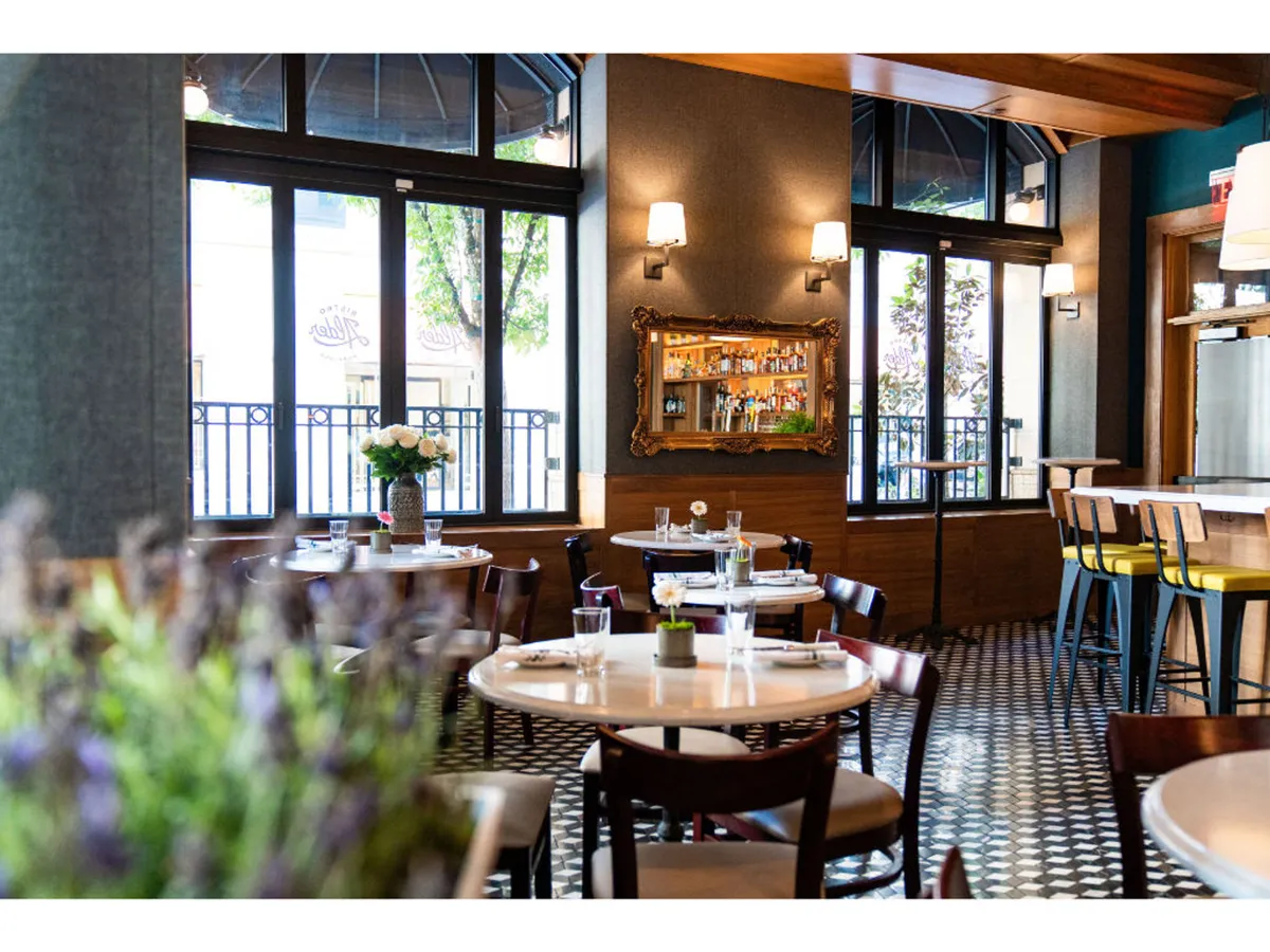 A cheerful space filled with natural light from large windows, floral centerpieces on tables, and a bar area reflecting bottles in a mirror. The casual yet inviting setting suggests a relaxed meal or happy hour with friends.