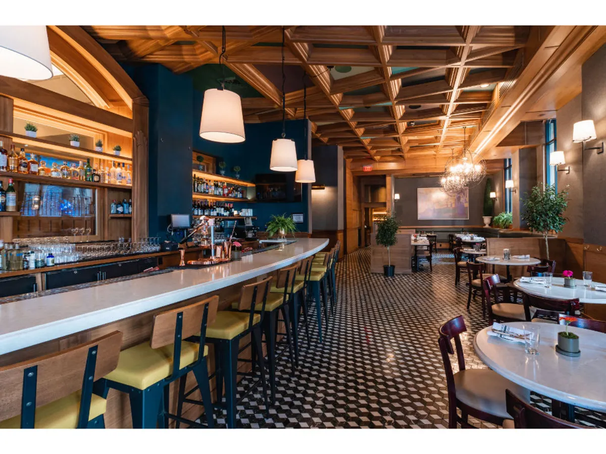 This image features a stylish bar counter set beneath a striking chandelier and a wooden ceiling, with inviting dining tables arranged nearby. The overall ambiance is sophisticated yet relaxed, suitable for both social dining and intimate gatherings. The space is well-lit and designed for comfort, with the bar area being a focal point.