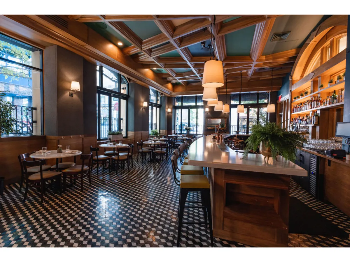 A stylish bar area anchors the foreground, with high-top tables and comfortable seating inviting guests to gather. The decorative ceiling and soft lighting create a welcoming yet sophisticated ambiance. Glassware and bottles line the shelves behind the bar, hinting at a menu of creative cocktails, while the open layout encourages social interaction.