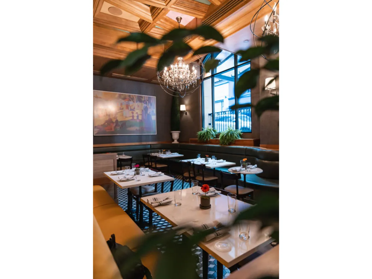 Tables set for dining are arranged beneath a modern chandelier, with contemporary artwork adorning the wall. Plant decorations add a touch of local greenery, while the overall setting suggests an artful, intimate dining experience.