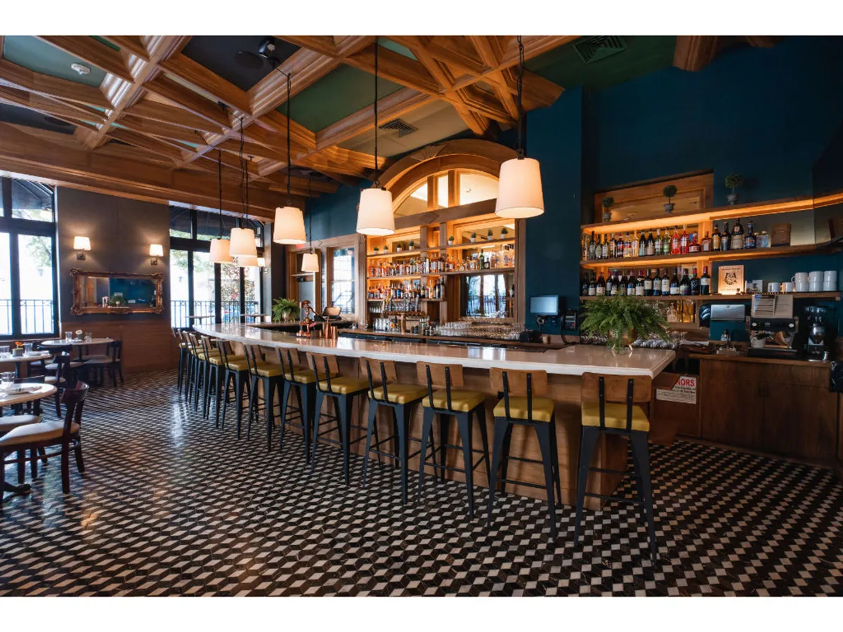 A sophisticated bar counter is lined with bottles on shelves, illuminated by hanging lights and framed by patterned floor tiles. The setting buzzes with energy as guests enjoy drinks and conversation, embodying the vibrant spirit of Bar Alder.