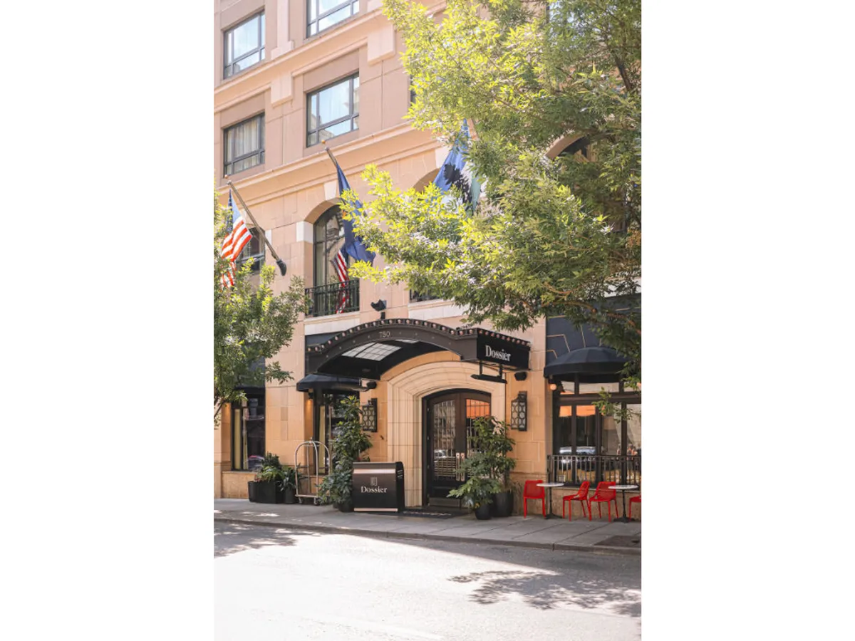 A lively exterior scene with an entrance awning, potted plants, flags, and vibrant red chairs that suggest a welcoming, active space for outdoor dining or socializing. The image conveys a sense of local culture and community activity, right at the doorstep.
